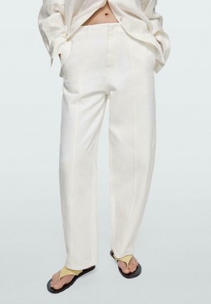 Persona che indossa pantaloni larghi color avorio, camicia aperta abbinata e sandali infradito beige, in piedi con le mani nelle tasche.