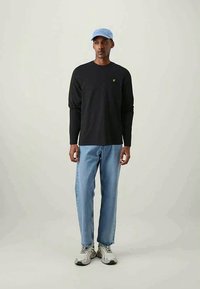 Camisa preta de mangas longas com logótipo amarelo, jeans de ganga azul, chapéu azul claro e sapatilhas cinzentas. O conjunto apresenta um corte casual e relaxado.