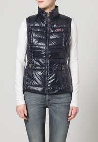Mujer vistiendo un chaleco acolchado navy brillante con parche de la bandera noruega, camisa blanca de mangas largas y jeans azules, de pie contra un fondo claro.