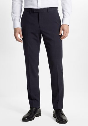 Man draagt op maat gemaakte donkerblauwe nette broek, een wit overhemd en zwarte leren nette schoenen, staand tegen een effen achtergrond.