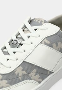 Baskets en toile grise avec accents en cuir blanc, présentant un motif subtil, des lacets plats et un détail de logo métallique sur la languette.