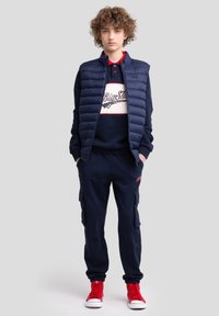 Gilet matelassé bleu marine sur un sweatshirt bleu marine avec un logo blanc et rouge. Pantalons de jogging assortis avec poches latérales ; baskets montantes rouges.