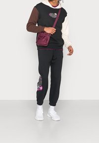 Černá a hnědá mikina s blokováním barev a krémovými detaily, doplněná černými tepláky s fialovým logem. Bílé tenisky a malá vínová kabelka.