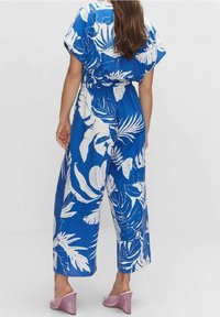 Modrý overal s bílými vzory listů, volný střih, elastický pás, krátké rukávy a široké nohavice. Nosený s obuví sv ětle fialové barvy.