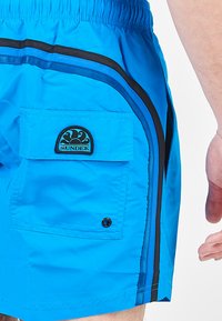 Costume da bagno in tessuto blu brillante con dettagli neri, dotato di tasca laterale e patch con logo. Vita elastica con cordino.