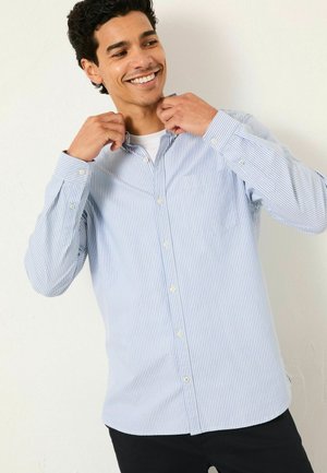 Hellblau gestreift, langärmliges Button-Up-Hemd. Verfügt über eine Brusttasche und weiße Knöpfe. Das Model justiert den Kragen und lächelt.
