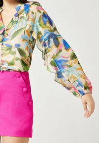 Blusa trasparente con vivaci motivi floreali in blu, verde e rosa; maniche lunghe e a sbuffo; abbinata a pantaloni corti fucsia con dettagli in bottone.