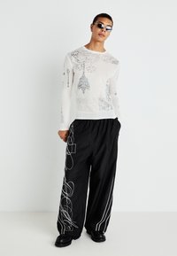 White long-sleeve knit shirt with graphic print, paired with loose black pants featuring white abstract patterns and chunky black shoes.