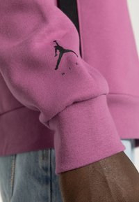 Primo piano di un braccio di pelle scura che indossa la manica di una felpa Nike rosa con il logo Jumpman nero, abbinato a jeans in denim blu chiaro.