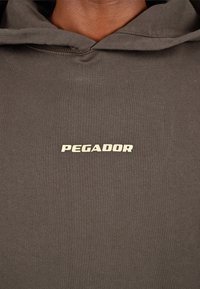 Σκούρο γκρι hoodie φτιαγμένο από μαλακό ύφασμα, με καγκουρό τσέπη και λογότυπο "PEGADOR" σε μπεζ χρώμα κεντραρισμένο στο στήθος.