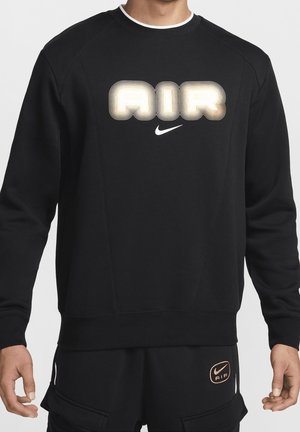 Osoba w czarnej bluzie Nike z dużym świecącym napisem "AIR" i logo Swoosh, w połączeniu z czarnymi spodniami z małym logo "AIR".