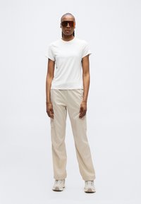 Γυναίκα με σκουρόχρωμο δέρμα, φορώντας ανακλαστικά γυαλιά ηλίου, λευκό κοντομάνικο πουκάμισο, μπεζ παντελόνι εργασίας cargo και ανοιχτόχρωμα αθλητικά παπούτσια, όρθια.