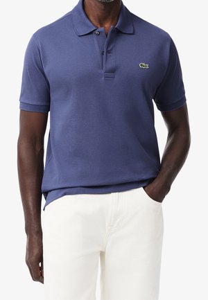 Mężczyzna w niebieskiej, krótkiej koszulce polo z małym zielonym logo aligatora na piersi i białych spodniach, z ręką w kieszeni.