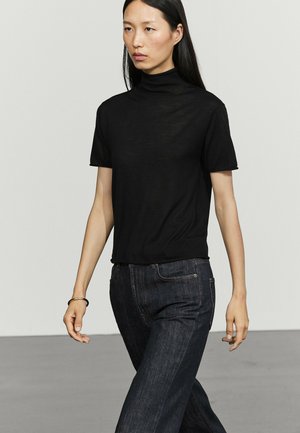 Donna con lunghi capelli neri che indossa un dolcevita nero a maniche corte e jeans blu scuro, cammina in un ambiente interno neutro.