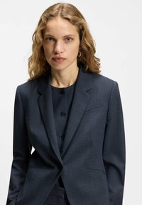 Blazer sartoriale blu navy in tessuto strutturato, con chiusura frontale a due bottoni, rever con intaglio e due tasche laterali.