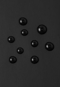 Dziewięć błyszczących czarnych kropli cieczy o różnych rozmiarach na ciemnej, fakturowanej powierzchni, ułożonych losowo.