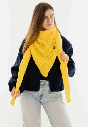 Yellow knitted triangular scarf with an open weave design, featuring long pointed ends and a decorative pin. Worn over a navy jacket.