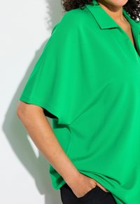 Grön oversized polo med krage, tillverkad av slät tyg; har korta ärmar och en avslappnad passform, parat med svarta byxor.
