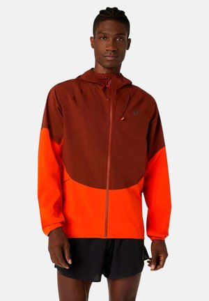 Homme portant une veste à capuche zippée bicolore marron et orange vif avec un short noir, debout sur un fond uni.