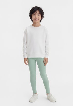 Kind steht, trägt ein weißes Sweatshirt, hellgrüne Leggings und weiße Turnschuhe, lächelt in die Kamera vor einem weißen Hintergrund.