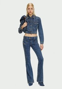 O tânără îmbrăcată cu o jachetă cropped texturată albastră și pantaloni evazați în tonuri corespunzătoare, ținând în spate o poșetă maro închis.