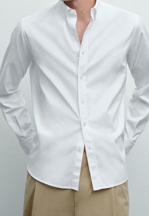 Homme portant une chemise blanche à manches longues déboutonnée sur un pantalon beige, debout les mains derrière le dos sur un fond clair uni.