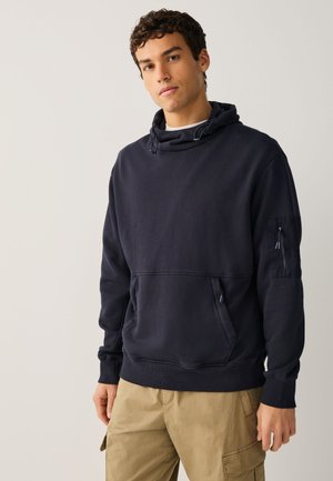 Jeune homme portant un sweat à capuche noir avec des poches sur le devant et les manches et un pantalon cargo beige, debout devant un fond clair uni.