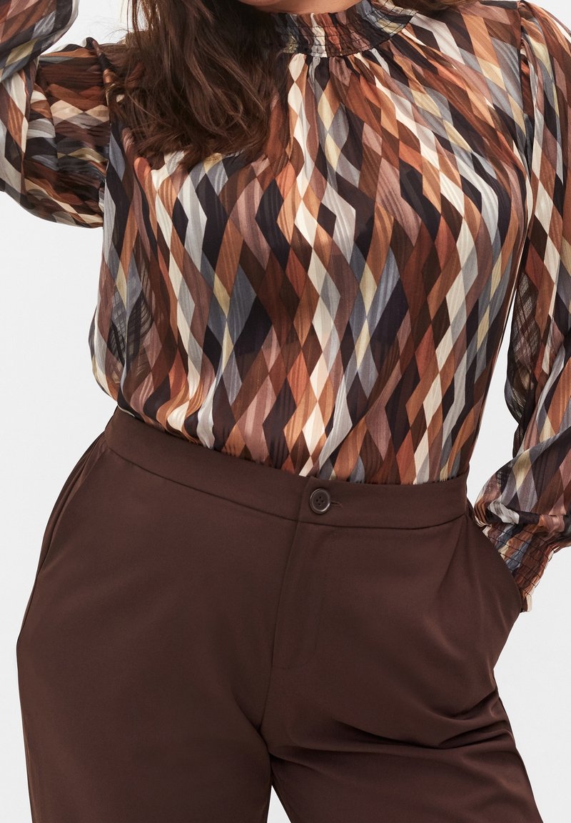 Femme portant un chemisier transparent à manches longues avec un motif géométrique marron, noir et beige, rentré dans un pantalon taille haute marron foncé.