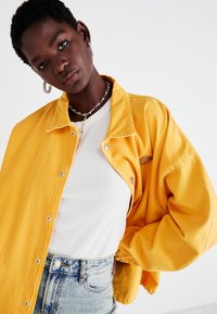 Gul denimjakke med avslappet passform, trykknapper og en subtil logo detalj. Båret over en ribbet hvit skjorte og slitte denimshorts.