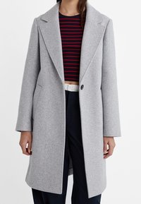 Grijs wollen overjas met een geknipte kraag, rechte snit en sluiting met één knop. Gedragen over een rood en marineblauw gestreept top.