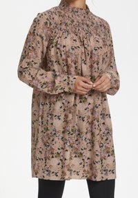 Tunique à motif floral en tissu doux et texturé, avec un col montant, des manches longues et une coupe décontractée. Les couleurs incluent le rose, le vert et le crème.