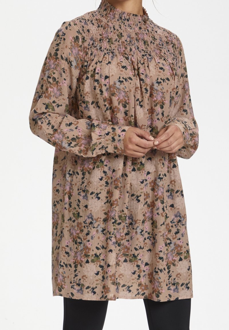 Tunique à motif floral en tissu doux et texturé, avec un col montant, des manches longues et une coupe décontractée. Les couleurs incluent le rose, le vert et le crème.