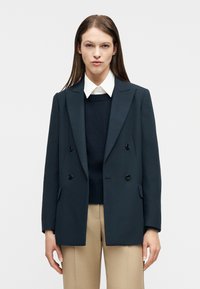 Γυναίκα με σκούρο διπλόμπουτο σακάκι, μπλε σκούρο πουλόβερ, λευκό πουκάμισο με γιακά και μπεζ παντελόνι, στέκεται μπροστά σε ένα απλό φόντο.