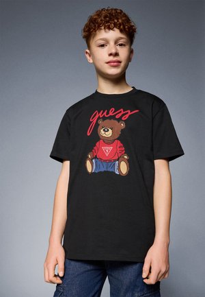 Schwarzes Baumwoll-T-Shirt mit einem Grafikdesign eines Bären in einem roten Pullover, mit dem Wort "guess" in Rot über dem Design. Kurze Ärmel, runder Hals.