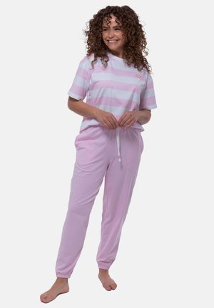 Femme souriante aux cheveux bouclés, vêtue d'un t-shirt rayé rose et blanc et d'un pantalon rose clair, debout pieds nus devant un fond uni.