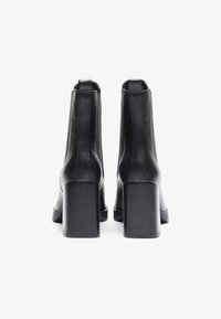 Bottines en cuir noir avec une texture lisse, talon carré, et des détails de couture minimalistes, vues de l'arrière.