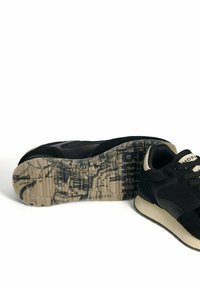 Zapatillas deportivas negras con parte superior de ante y malla, acentos beige y una suela de goma con patrón que presenta diseños impresos.