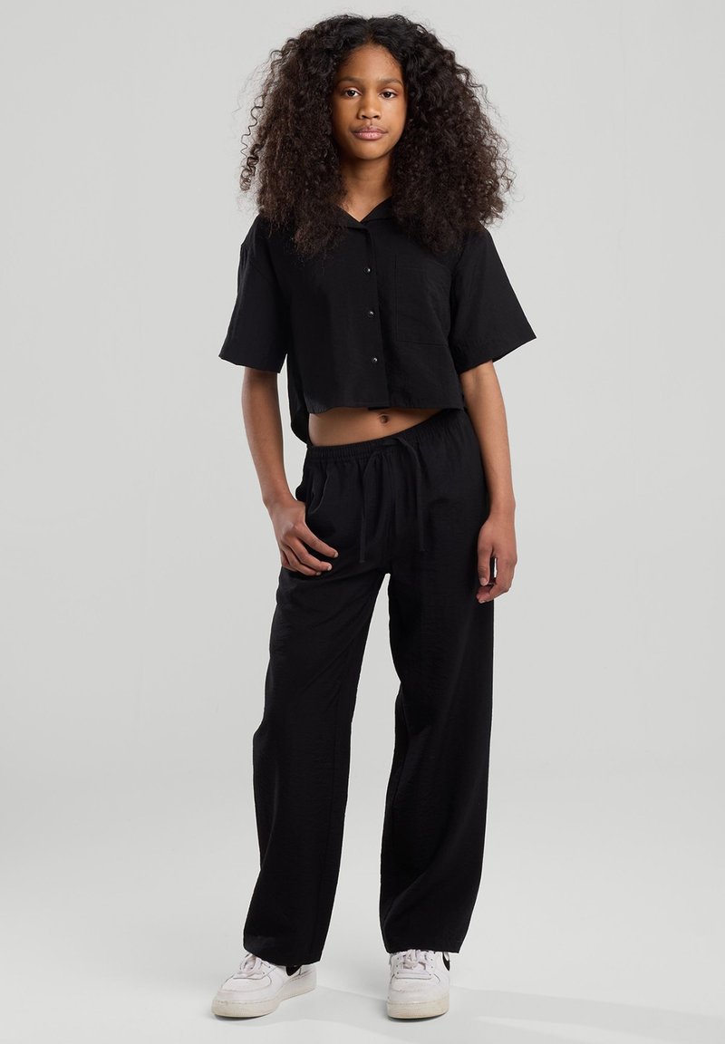 Mustat lyhythihaiset napitettava crop-paita, joka on yhdistetty löysiin mustiin housuihin. Molemmat vaatteet ovat pehmeää, teksturoitua kangasta. Valkoiset tennarit.