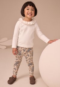 Maglia bianca a maniche lunghe con scollatura arricciata, leggings a motivo floreale in beige, blu e marrone, e scarpe grigie con fibbie. Sfondo uniforme.