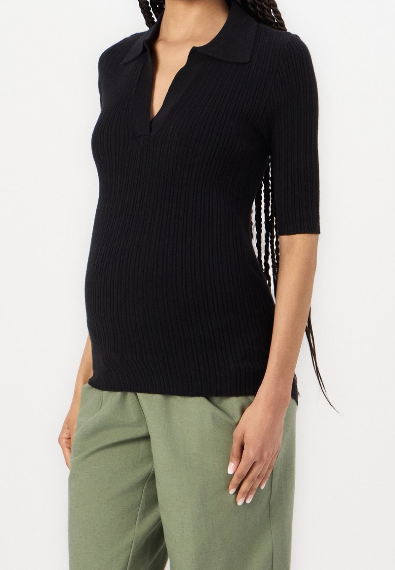 Femme portant un polo noir côtelé à manches courtes et un pantalon ample vert olive, avec de longs cheveux tressés sur l'épaule.