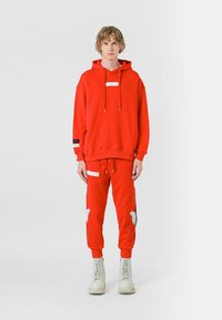 Sudadera roja con capucha y joggers a juego, hechos de felpa, con un parche blanco y diseños bordados. El modelo lleva botas blancas con cordones.