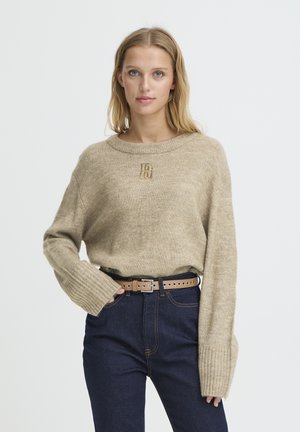 Pull beige en maille avec un col large et arrondi, des poignets côtelés et un logo texturé sur la poitrine, porté avec un jean en denim foncé et une ceinture marron.