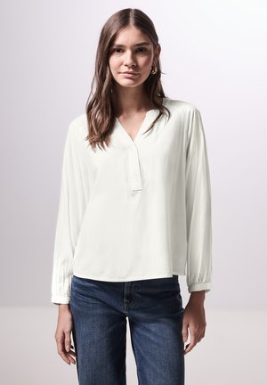 Witte blouse met lange mouwen, V-hals en knoopsluiting; gemaakt van zacht, glad materiaal en heeft een losse pasvorm. Gepaard met donkerblauwe jeans.