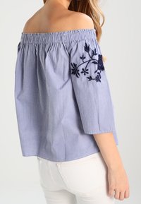 Blouse dégagée en bleu et blanc à rayures, ornée d'une broderie florale marine sur la manche, avec une coupe décontractée et un décolleté élastique.