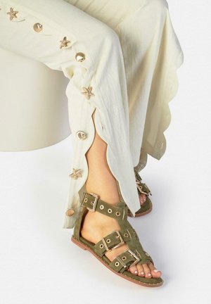 Benenden in crèmekleurige broek met zeesterren- en schelpknopen en olijfgroene sandalen met meerdere gespen en metalen ringen.