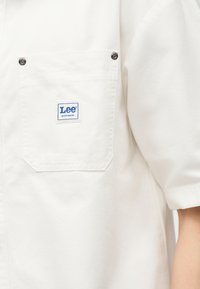 Λευκό πουκάμισο εργασίας με κοντό μανίκι και μπροστινή τσέπη ασφαλισμένη με δύο μεταλλικά καρφιά, με ένα μικρό μπλε λογότυπο Lee Workwear.