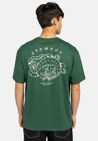 T-shirt in cotone verde con un ampio grafico bianco di un serpente sul retro e la dicitura "ELEMENT" sopra di esso. Maniche corte.