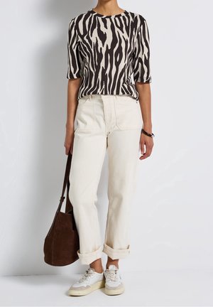 Person trägt ein schwarz-weißes Zebramuster-Top, hochgekrempelte cremefarbene Hose, weiße Sneakers und hält eine braune Wildleder-Schultertasche.