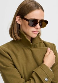 Cappotto di lana verde oliva con colletto alto, caratterizzato da un dettaglio con bottone sul polsino; modello con occhiali da sole marroni oversize, capelli lunghi fino alle spalle.