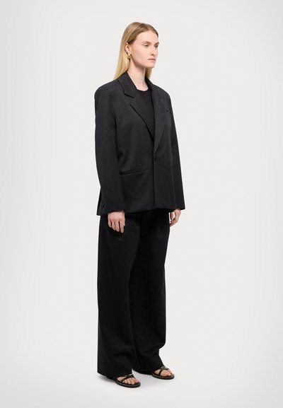 Blazer noir ajusté avec une silhouette structurée, associé à un pantalon noir à jambes larges. Porté sur un haut noir, accentué par des sandales plates.